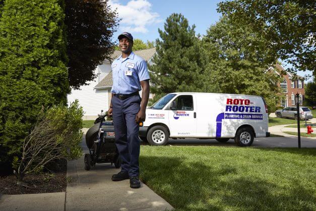 Plumbing technician arriving at customer residence with drain cleaning machine in tow, company-branded van sitting in driveway behind
